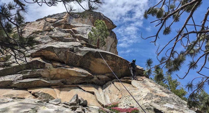 From the ground looking up, a person wearing safety gear is secured by ropes as they stand on the ledge of a rocky cliff. 